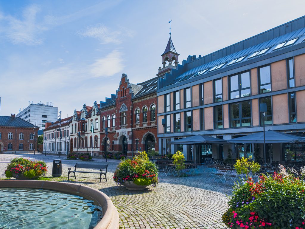 Plac w centrum Kristiansand z zabytkową zabudową, fontanną i kolorowymi kwiatami.