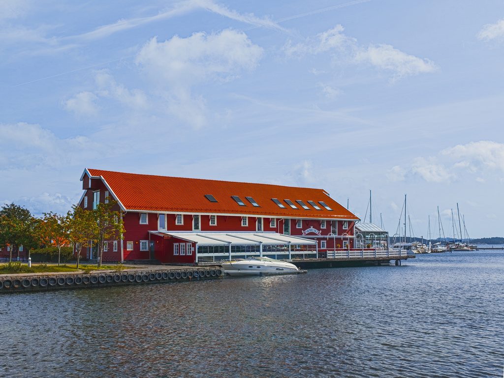 Czerwony drewniany budynek z pomarańczowym dachem nad wodą w Kristiansand, z przystanią jachtową i żaglówkami w tle.