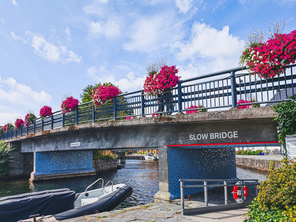 Most SLOW BRIDGE w Kristiansand ozdobiony różowymi kwiatami, nad kanałem z zacumowanymi łodziami.