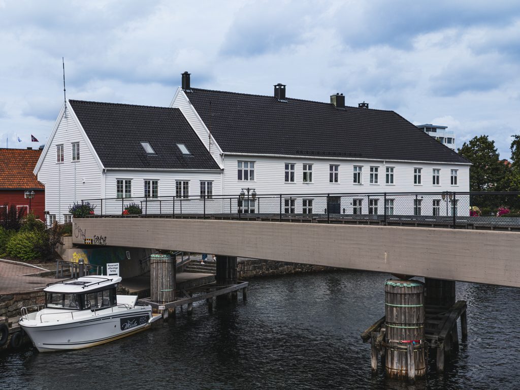 Biały drewniany budynek nad kanałem w Kristiansand, z mostem i łodzią zacumowaną przy nabrzeżu.
