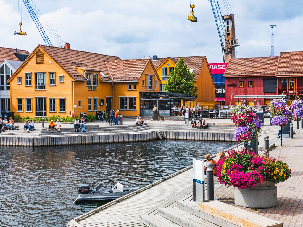 Kolorowe drewniane budynki przy nabrzeżu Fiskebrygga w Kristiansand, z kwiatami, restauracjami i odpoczywającymi nad wodą ludźmi.