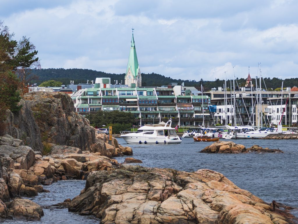 Skaliste wybrzeże Kristiansand w Norwegii, z widokiem na port jachtowy, zacumowane łodzie i zabudowę miasta z wieżą kościelną w tle.